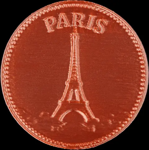 Detalhe macro de uma moeda decorativa de Paris com a Torre Eiffel em relevo, mostrando o acabamento cor de cobre e a borda decorada.